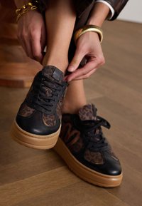Schwarze und braune Sneakers mit einem Leopardenmuster, dicken Sohlen und schwarzen Schnürsenkeln. Das strukturierte Material umfasst Leder- und Stoffakzente.