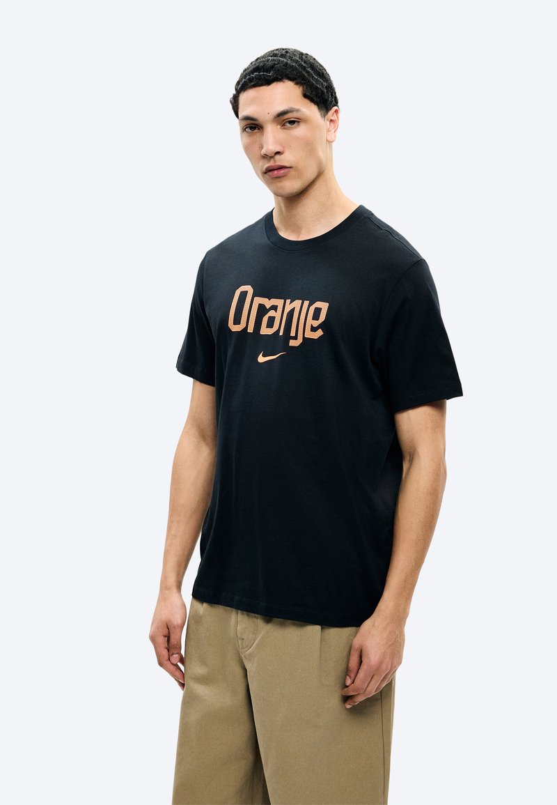 Νέος άνδρας που φοράει μαύρο T-shirt με τη λέξη "Orange" και το λογότυπο της Nike σε πορτοκαλί, συνδυασμένο με μπεζ παντελόνια, στέκεται μπροστά σε απλό λευκό φόντο.
