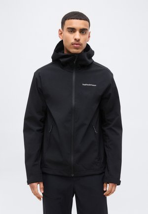 Νεαρός άνδρας που φοράει μαύρο μπουφάν με κουκούλα, φερμουάρ στο μπροστινό μέρος και τσέπες, στέκεται μπροστά σε απλό ανοιχτό γκρι φόντο.