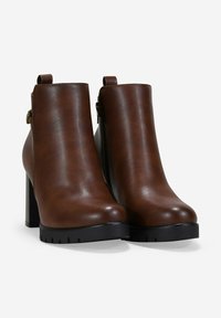 Botas de cuero marrón hasta el tobillo con tacones altos y gruesos, suelas negras de tracción y cremalleras laterales sobre un fondo blanco.