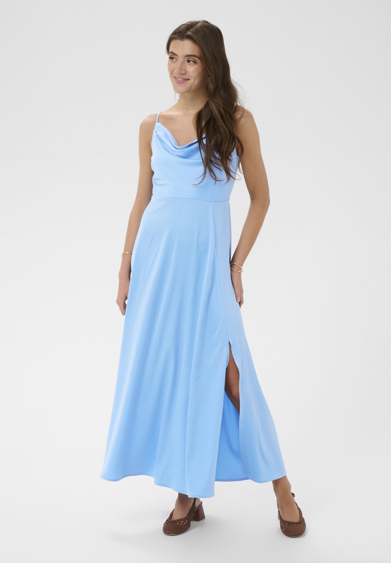 Jeune femme portant une robe longue bleu clair sans manches avec une fente latérale et des chaussures marron tressées à talon bloc, souriante et regardant sur le côté.
