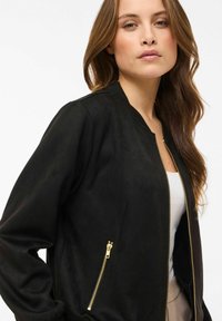 Juoda bombonešio striukė iš minkštos medžiagos, su ribiniu apykakliu, ilgomis rankovėmis ir auksinėmis kišenėmis su užtrauktukais priekyje. Lygi tekstūra, minimalistinis dizainas.