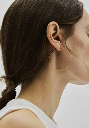 Barna hajú nő, aki alacsonyan összefogott copfját viseli, nagy aranykarika fülbevalót és fehér, ujjatlan felsőt, jobbra néz.