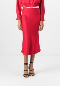 Jupe midi en satin rouge avec une texture lisse, légèrement évasée à l'ourlet, associée à des sandales à talons hauts noires à lanières.