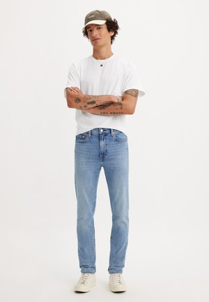 Jeans azzurri chiari a taglio slim, indossati con una t-shirt bianca e un cappellino grigio. Caratteristiche includono tatuaggi visibili e sneakers beige.