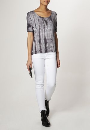 Kobieta ubrana w szarą, krótko-rękawową bluzkę z efektem tie-dye, białe obcisłe dżinsy i czarne botki do kostek, trzymająca w prawej ręce czarną kopertówkę.