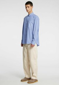 Zils un balts svītrains krekls ar pogām aiztaisāmu apkakli, sapārots ar bēšām biksēm un brūnām kurpēm. Redzama krūšu kabata un brīvs piegriezums.