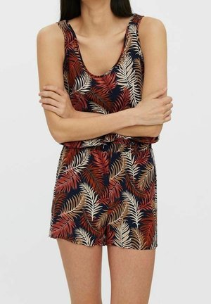 Bezrekawowy kombinezon z granatowym tłem i czerwonym, beżowym oraz brązowym tropikalnym wzorem liści, noszony przez osobę z założonymi ramionami.