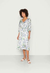 Vestido floral em tecido leve com decote em V, mangas curtas e cintura com laço. As cores incluem azul, verde e branco. Estilizado com sapatos brancos.