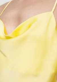 Rumena satenasta majica z ovratnim izrezom in tankimi nastavljivimi naramniki, s sijočo, gladko in mehko teksturo.