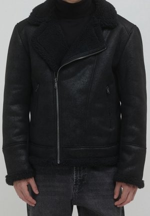 Mand iført sort shearlingjakke med asymmetrisk lynlås og mørke jeans, stående mod en ensartet lys baggrund.