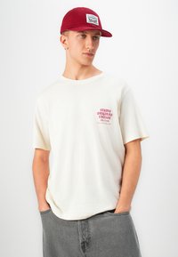 Joven vestido con una camiseta blanca "Studio Originals Edition", gorra roja y jeans grises, de pie con las manos en los bolsillos, mirando de lado.