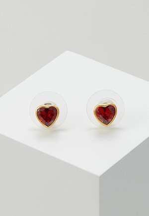 Pair of small heart-shaped red gemstone stud earrings with gold trim on clear backing, displayed on white angled surface.
