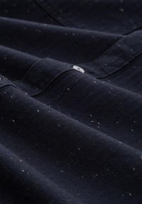 Dunkelblauter Stoff mit strukturiertem Fischgrätmuster, versehen mit verstreuten weißen und roten Sprenkeln sowie einem kleinen Knopfdetail.