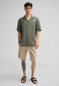 Chemise à manches courtes vert olive avec des boutons et une poche avant, associée à un short beige mi-cuisse et des sandales noires. Texture du tissu lisse.