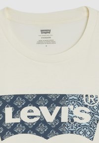 Kremfarget bomull T-skjorte med et stort blått og hvitt Levi's-logo med blomster mønster i bakgrunnen og rund hals. Størrelse M.