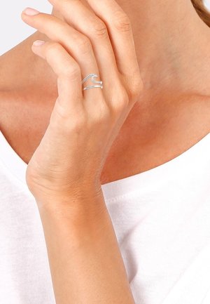 Main avec une bague argentée en forme de vague portée à l'annulaire, touchant le visage près du menton, portant une chemise blanche, teint clair.