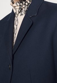 Námorně modrý blazer s dvěma knoflíky, klopovým límcem a jemnými stehy. Podsazený s košilí s vzorovaným límcem v světlých tónech.