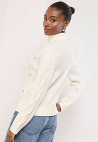 Fehér, kötött pulóver magas gallérral, a ujjain texturált mintázattal és díszgombokkal az elülső részen.