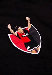 Zwarte stof bevat een geborduurd embleem van een voetballer in een rood en zwart tenue, met opgestoken armen. De schildvorm heeft een witte rand en bevat het nummer "10."