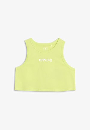 Dziewczyna z długimi ciemnymi włosami, ubrana w limonkową, bezrękawnikową bluzkę typu crop top, szare szorty z elastycznym pasem, białe skarpetki i fioletowe trampki, stoi z rękami w kieszeniach.