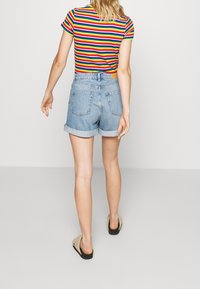 Camisa de manga corta a rayas de múltiples colores, pantalones cortos de mezclilla de talle alto, con los puños enrollados y espalda descubierta. Sandalias con suelas negras.
