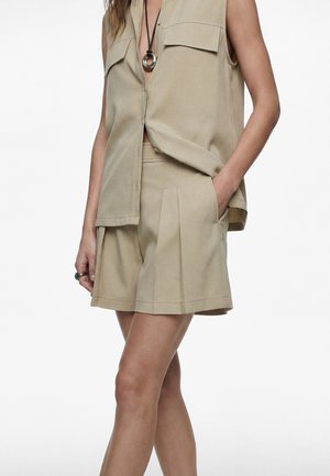 Kvinde iført beige ærmeløs knap-op skjorte og matchende plisserede shorts, med hånden i lommen og sølvvedhængs halskæde.