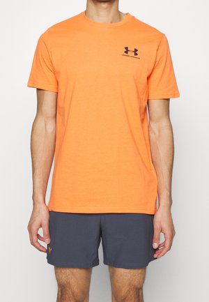 Mężczyzna w jaskrawo pomarańczowej koszulce Under Armour z krótkim rękawem i ciemnoszarych szortach sportowych stoi na białym tle.