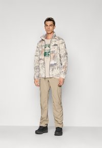 Mann iført en beige grafisk t-skjorte, lysmønstret jakke med glidelås, khaki cargo-bukser, svarte tursko, og solbriller på hodet.