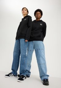Two people standing side by side wearing black hoodies with white heart logos and loose blue jeans against a plain white background.