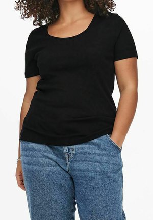 Kobieta o kręconych włosach, ubrana w czarną bluzkę z krótkim rękawem i niebieskie dżinsy, z jedną ręką w kieszeni na białym tle.