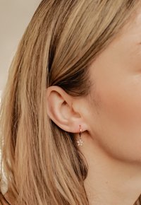 Złoty kolczyk w formie kółka z wiszącym urokiem w kształcie gwiazdki, ozdobionym przezroczystymi kamieniami, przedstawiony na tle długich, prostych włosów i ucha.