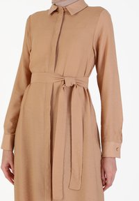 Beige långärmad klänning med en krage och ett tygbälte knutet i midjan, bärs av en person med avslappnade händer vid sidorna.