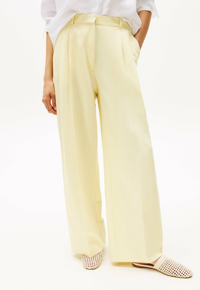 Pantalones de tiro ancho en color amarillo claro con una textura suave, diseño de frente plisado, combinados con zapatos de lona sin cordones. Modelo de pie.