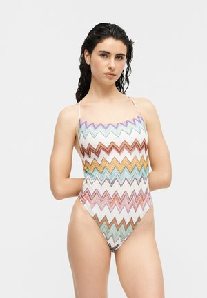 Γυναίκα με σκούρα, βρεγμένα μαλλιά, που φοράει ολόσωμο μαγιό με πολύχρωμο σχέδιο ζιγκ-ζαγκ και λεπτές τιράντες στους ώμους, πάνω σε φωτεινό φόντο.