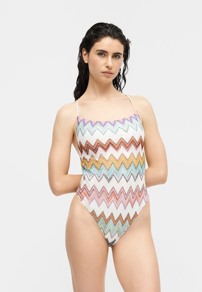 Femme aux cheveux noirs mouillés portant un maillot de bain une pièce multicolore à motif en zigzag avec des bretelles fines sur un fond clair.