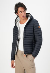Man in een marineblauwe gewatteerde jas met capuchon over een wit T-shirt en beige broek, kijkend naar links met een hand in zijn zak.