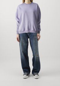 Sudadera lila con hombros caídos, combinada con jeans azules desgastados. El modelo lleva zapatillas negras con suelas blancas.