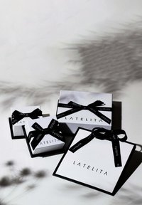 Tre scatole regalo bianche con nastro nero caratterizzanti il logo "LATELITA". Di diverse dimensioni, con forma quadrata e superficie liscia. Ombre visibili al di sotto.