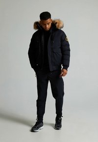 Svart boblejakke med hette av kunstpels, frontglidelås og sidelommer. Kombinert med mørke bukser og svarte treningssko.