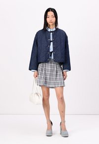 Marineblauwe gewatteerde bomberjack met drukknoopsluiting, lichtblauwe blouse, geruite plissérok, grijze plateauhakken en witte handtas.