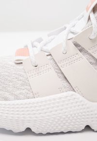 Kevyt harmaa urheilukenkä, jossa on teksturoitu neulottu päällinen, valkoinen kumipohja ja hillityt vaaleanpunaiset yksityiskohdat remmeissä ja kielen alueella.