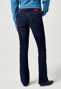 Jeans de campana azul oscuro con un diseño clásico de cinco bolsillos. Presentan un parche de cuero marrón en la cinturilla y un patrón de costura sutil en los bolsillos.