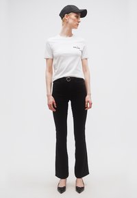 Άτομο που φοράει λευκό μπλουζάκι με τη λέξη "AMOUR", μαύρο καμπάνα παντελόνι με ζώνη, μαύρα μυτερά παπούτσια και μαύρο καπέλο, στέκεται μπροστά σε λευκό φόντο.