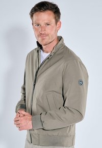 Lightweight olive green bomber jacket with a zip front, side pockets, ribbed cuffs, and a logo patch on the left sleeve. Smooth texture.