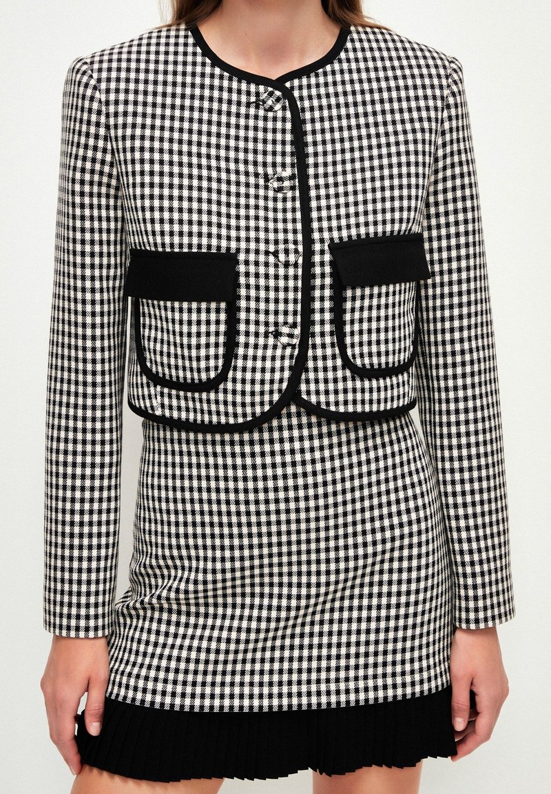 Veste courte à carreaux noir et blanc avec poches à rabat bordées de noir, portée avec une mini-jupe assortie à l'ourlet plissé noir.