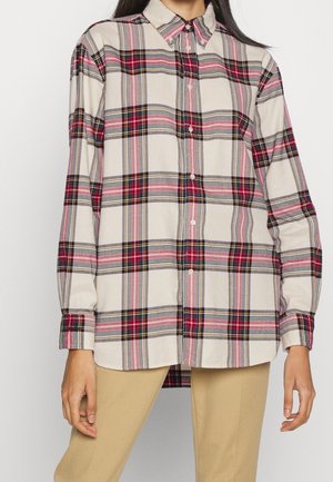 Femme portant une chemise boutonnée à manches longues avec un motif à carreaux rouge, noir, rose et beige, associée à un pantalon beige.