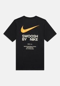 Melnā Nike T-kreklā ar lielu oranžu Swoosh logotipu un baltu tekstu "SWOOSH BY NIKE", kā arī uzņēmuma adresi uz aizmugures.