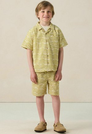 Kurze Ärmel Knopfhemd und Shorts-Set in blassem Gelb mit wellenförmigen weißen Mustern. Kombiniert mit hellbraunen, weichen Schuhen.