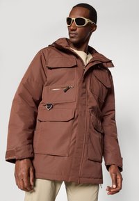 Veste isolée marron avec plusieurs poches, une fermeture éclair à l'avant, une capuche ajustable et des poignets texturés. Le modèle porte un pantalon de couleur claire et des lunettes de soleil.